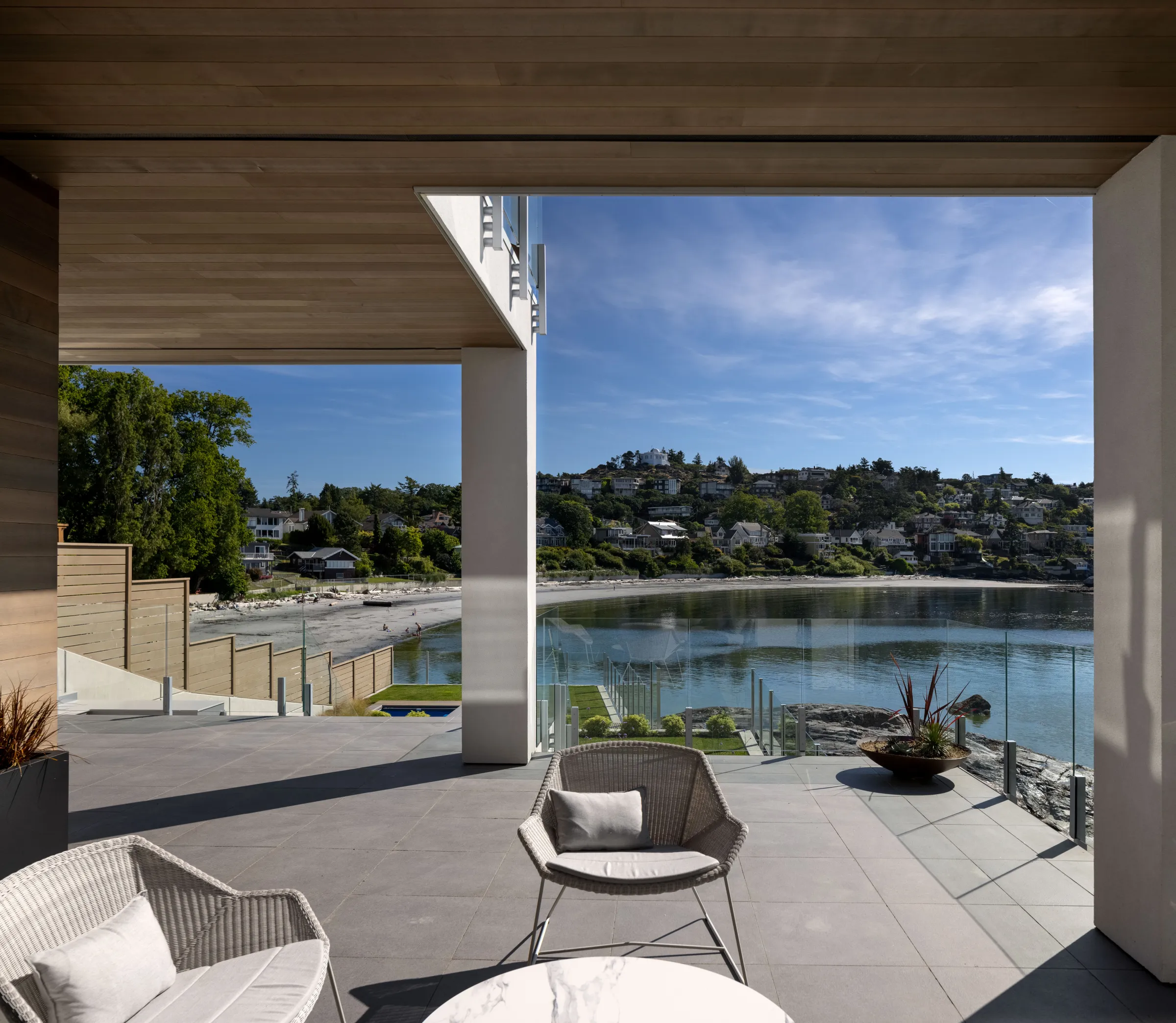Covered terrace under a cedar soffit, wicker chairs on grey porcelain pavers above a sheltered beach