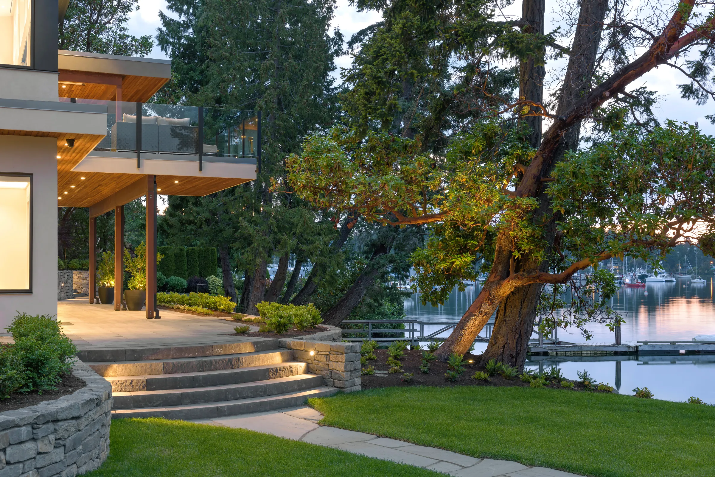 Cantilevered deck on timber posts lit at dusk, stone steps down to lawn, arbutus and moored boats beyond