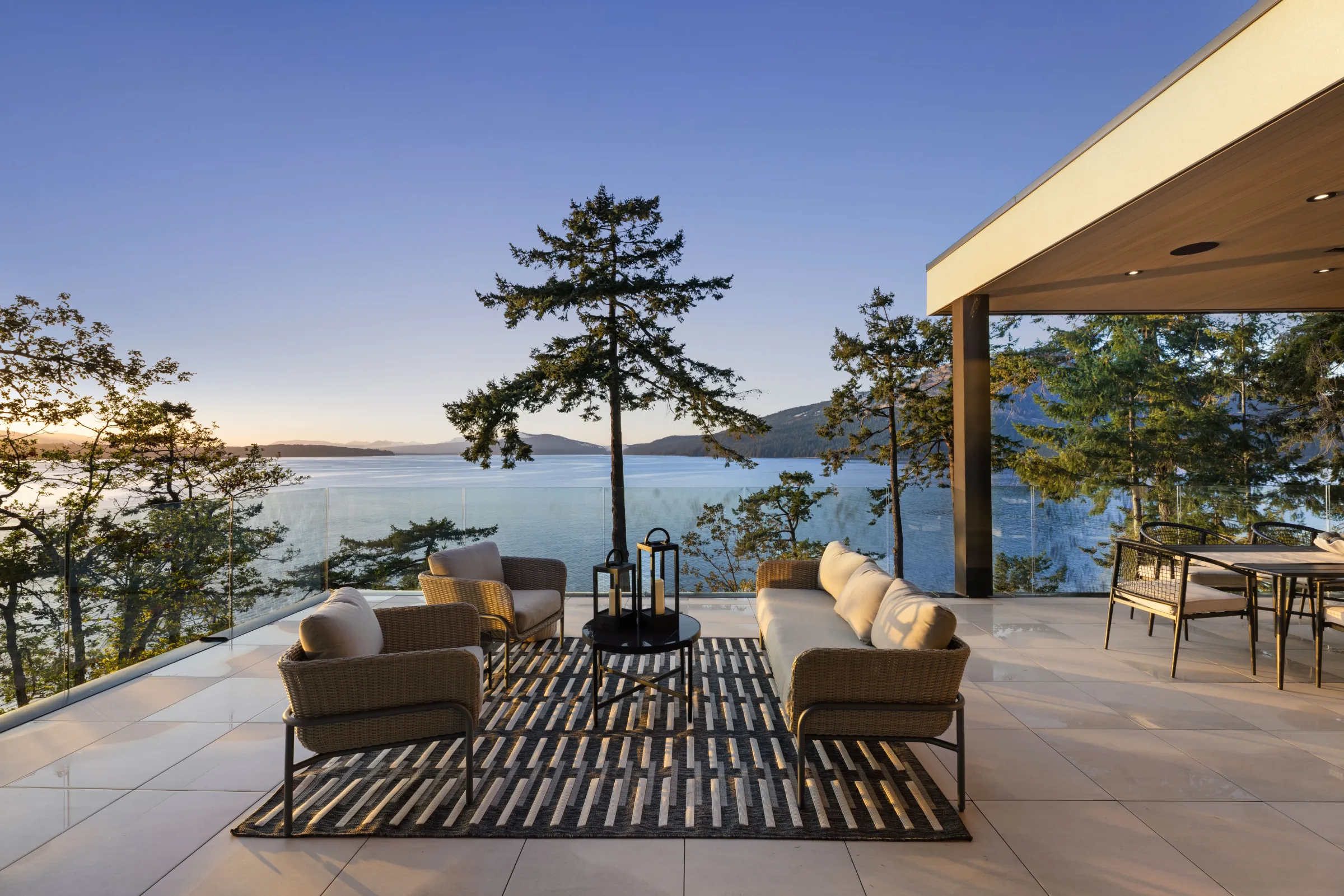 Upper terrace at dusk behind frameless glass, wicker seating on a striped rug, a single fir against the strait