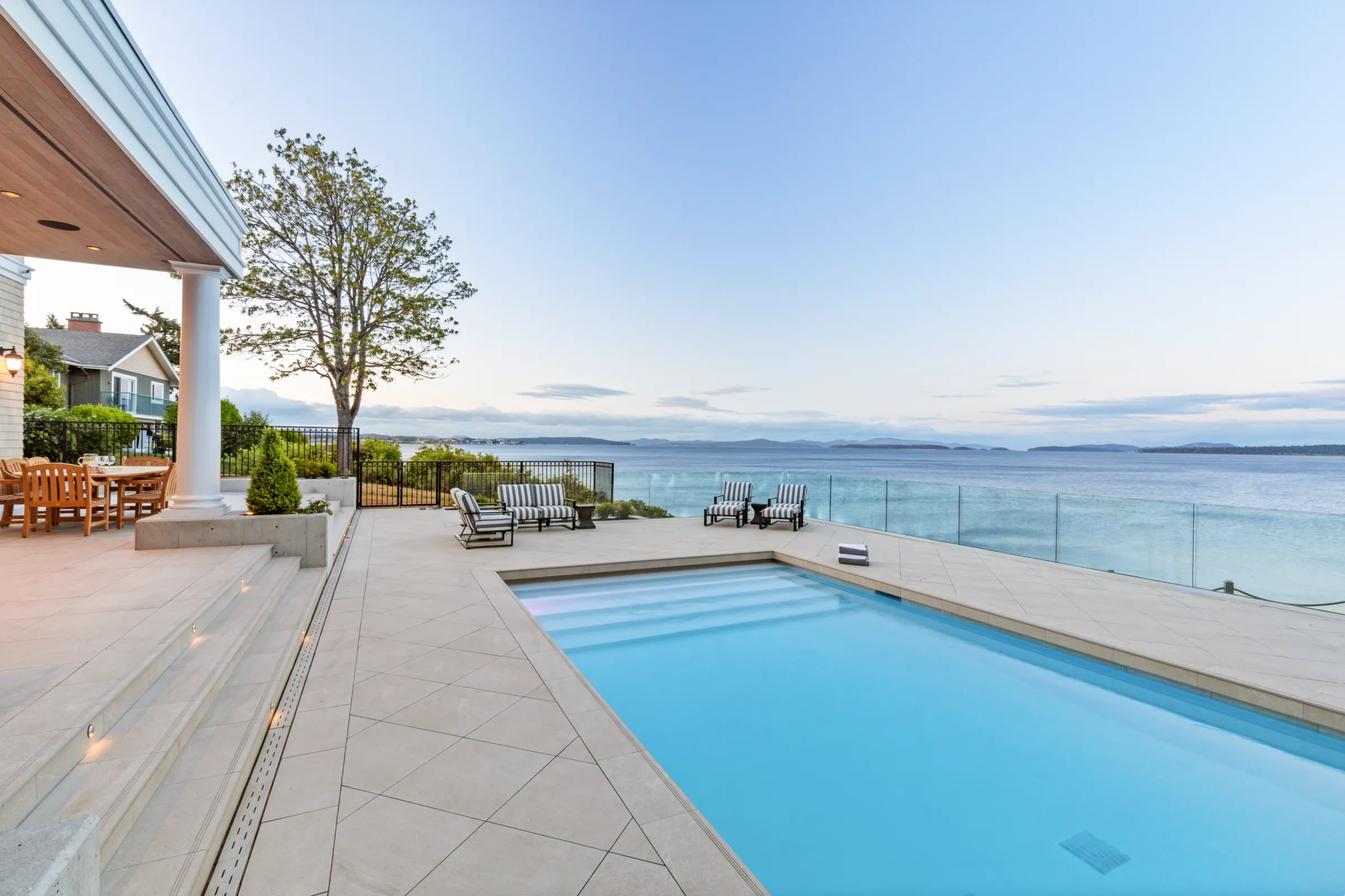 Lap pool on a stone deck beside a columned porch, glass guardrail open to the water and distant islands