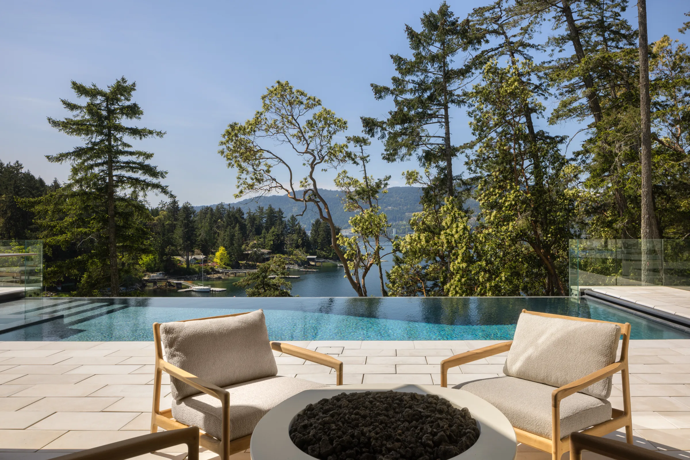 Infinity pool and round fire table with teak chairs, arbutus and firs above a bay of moored sailboats